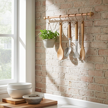 Load image into Gallery viewer, Wooden spoons hanging on a copper rod against a brick wall with a window in the background.